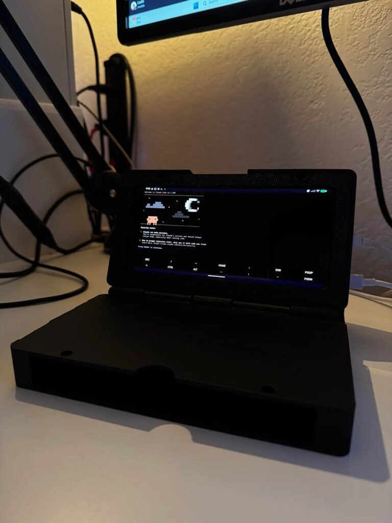 Um computador portátil com uma tela monocromática exibindo uma interface de arte ASCII está sobre uma mesa, ao lado de um braço ajustável para microfone.