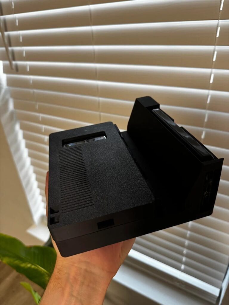 Uma pessoa segura um dock preto para jogos, sem marca, com padrões de ventilação e encaixes de portas visíveis, perto de uma janela com persianas.